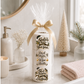 A gift-wrapped stack of three toilet paper rolls sits on a bathroom counter. The rolls have holiday-themed printed labels reading "Santa's Been Too Busy For Your Turn," "Deck Halls Wipe Balls," and "Shit Happens When Christmas.
